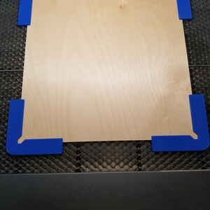 May include: A piece of plywood with blue corner clamps. The plywood is on a black honeycomb surface.
