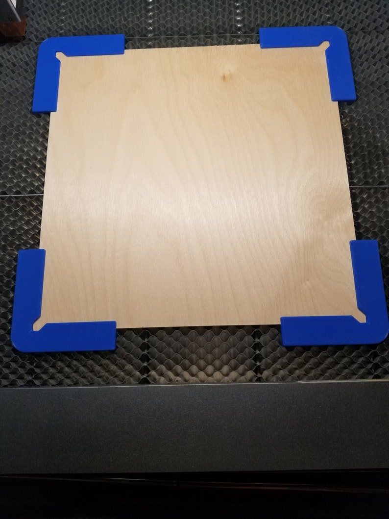 May include: A piece of plywood with blue corner clamps. The plywood is on a black honeycomb surface.