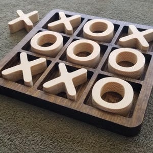 May include: A wooden tic-tac-toe game set featuring a dark brown square board with a grid of nine recessed squares. The set includes light-colored wooden game pieces shaped as X and O symbols, with several pieces placed inside the grid.