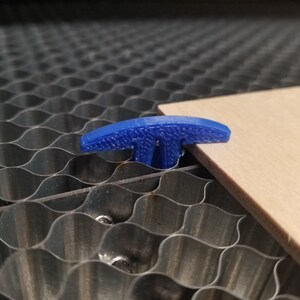 Pu&ograve; includere: Un dispositivo di fissaggio a forma di T in plastica blu viene utilizzato per tenere un pezzo di legno in posizione su un tappetino da taglio a nido d'ape.