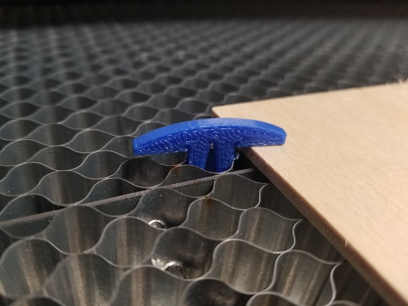 Pu&ograve; includere: Un dispositivo di fissaggio a forma di T in plastica blu viene utilizzato per tenere un pezzo di legno in posizione su un tappetino da taglio a nido d'ape.