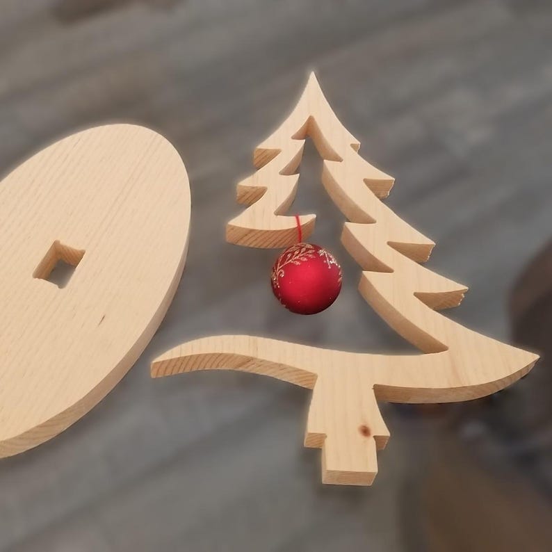 May include: A light-colored wooden cutout in the shape of a stylized Christmas tree with a small red spherical ornament hanging from the center. Beside the tree is a circular wooden base featuring a square cutout in the middle for assembly.