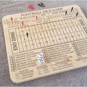 May include: A wooden football dice game board featuring a printed field layout with yard markers, scoreboards, and play outcome charts. The board includes red, white, and black pegs for tracking game progress, along with two white six-sided dice.