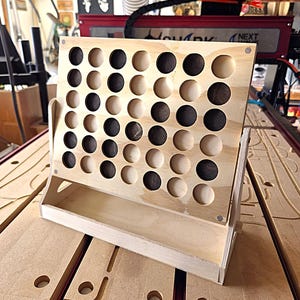 Puede incluir: Un tablero de juego de cuatro en raya de madera con un marco de madera contrachapada clara y una rejilla de recortes circulares. El tablero contiene piezas de juego circulares de madera, alternando tonos oscuros y claros, en las ranuras.