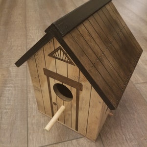 May include: A wooden birdhouse with a brown roof and a small perch. The birdhouse has a small door with a circular opening and a decorative design on the front.