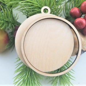 May include: A wooden Christmas ornament with a circular design. The ornament has a round, flat wooden base with a circular frame around it. The ornament is hanging from a small loop at the top. The ornament is surrounded by green pine needles and red berries.