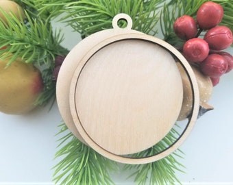 May include: A wooden Christmas ornament with a circular design. The ornament has a round, flat wooden base with a circular frame around it. The ornament is hanging from a small loop at the top. The ornament is surrounded by green pine needles and red berries.