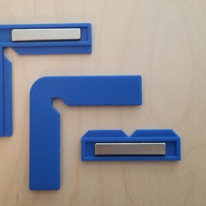 May include: Three blue plastic corner clamps with silver metal magnets. The clamps are designed to hold pieces of wood together at a 90-degree angle.