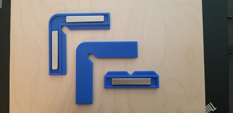 May include: Three blue plastic corner clamps with silver metal magnets. The clamps are designed to hold pieces of wood together at a 90-degree angle.