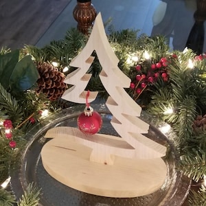 May include: A light-colored wooden Christmas tree cutout standing on an oval wooden base. A single small, round, red ornament with gold-colored floral detailing hangs from a branch of the tree by a thin red string.