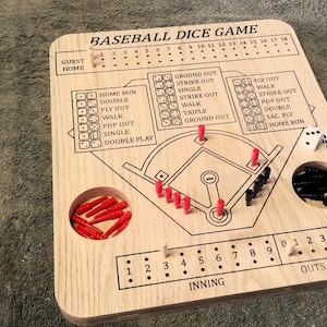 May include: A light-colored wooden baseball dice game board featuring a printed field diagram, score tracking sections for guest and home teams, inning and out counters, and holes for pegs. Includes red and black plastic pegs and white dice.