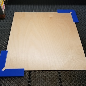 May include: A light brown piece of wood with a smooth surface is positioned on a black honeycomb surface. Two blue corner clamps are holding the wood in place.