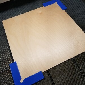 May include: A piece of plywood is secured to a laser cutting bed using blue plastic corner clamps.