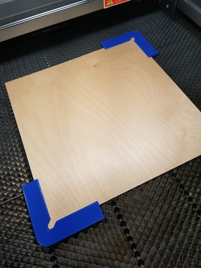 May include: A piece of plywood is secured to a laser cutting bed using blue plastic corner clamps.