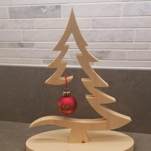 May include: A light-colored wooden Christmas tree cutout standing on a circular wooden base. A single round red ornament with a gold floral pattern hangs from a branch by a thin red string, centered within the open space of the tree structure.