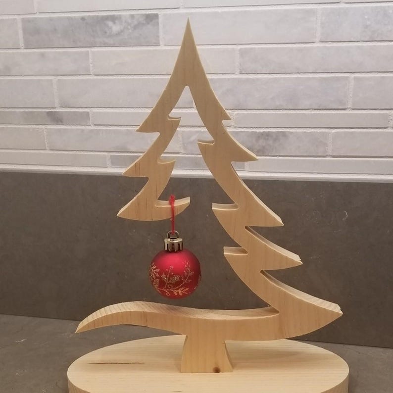 May include: A light-colored wooden Christmas tree cutout standing on a circular wooden base. A single round red ornament with a gold floral pattern hangs from a branch by a thin red string, centered within the open space of the tree structure.