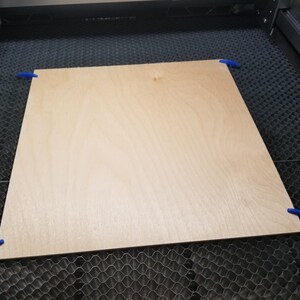 Pu&ograve; includere: Un pezzo di legno marrone chiaro &egrave; fissato a una superficie a griglia nera con clip blu. Il legno &egrave; pronto per essere tagliato da una taglierina laser.