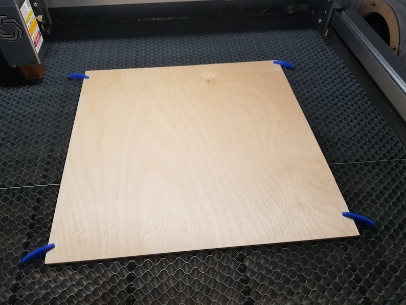 Pu&ograve; includere: Un pezzo di legno marrone chiaro &egrave; fissato a una superficie a griglia nera con clip blu. Il legno &egrave; pronto per essere tagliato da una taglierina laser.