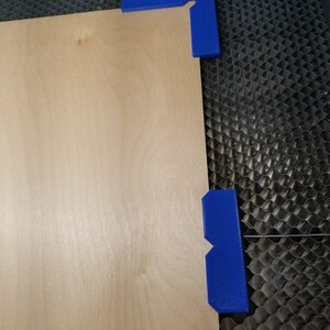 May include: A piece of light brown plywood is secured to a black honeycomb surface with two blue plastic corner clamps.