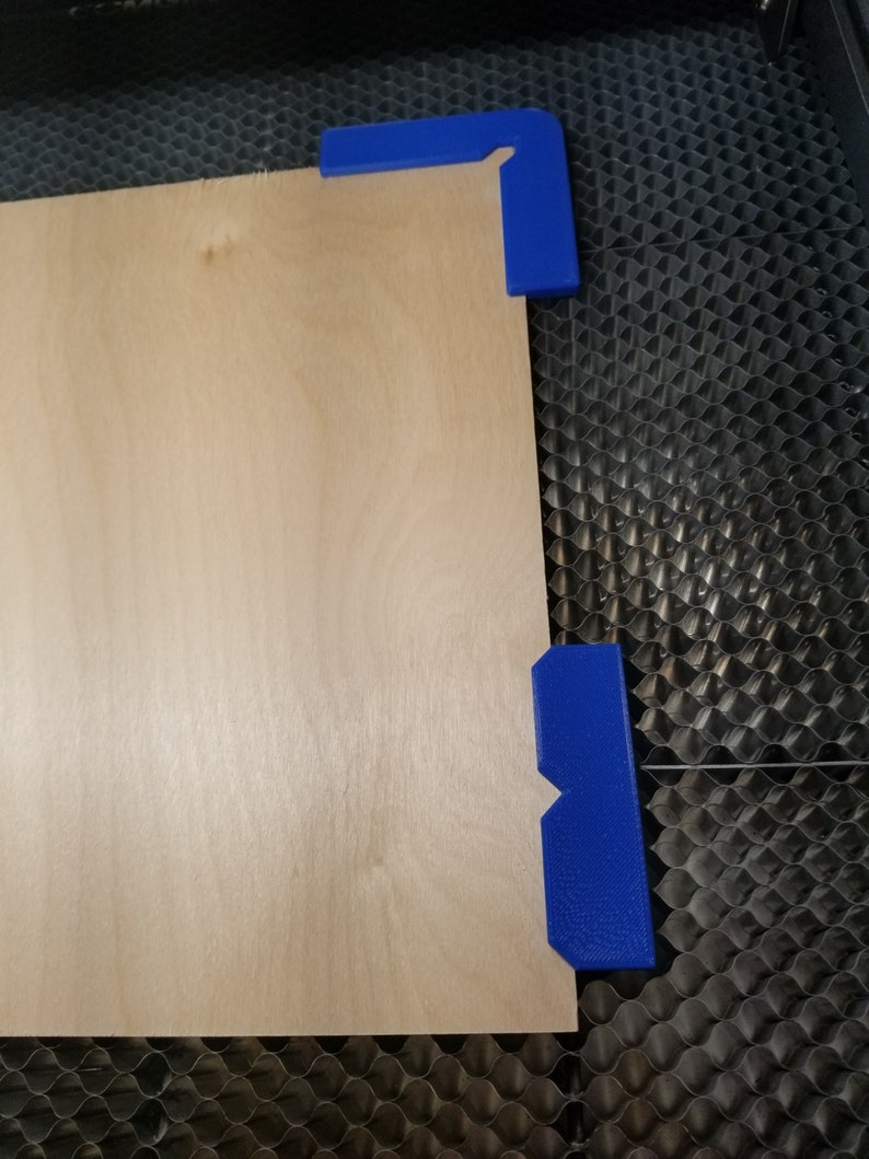 May include: A piece of light brown plywood is secured to a black honeycomb surface with two blue plastic corner clamps.
