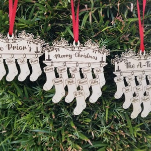 May include: Three wooden Christmas ornaments with red ribbons. Each ornament features a decorative top with text and a series of hanging stockings with names. The texts read "The Prior's", "Merry Christmas", and "The A-Team".