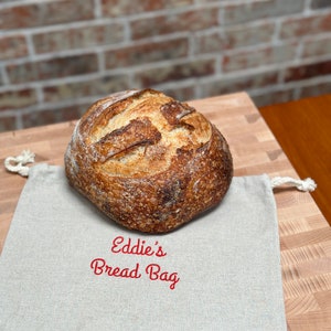 May include: A light brown sourdough bread loaf sits on a beige linen bread bag with the text "Eddie's Bread Bag" embroidered in red.