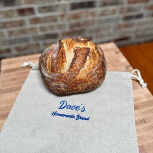 May include: A freshly baked loaf of sourdough bread sits on a beige linen bread bag with the text "Dave's Homemade Bread" embroidered in blue.