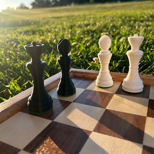 May include: A wooden chessboard with black and white squares. There are three chess pieces on the board: a black queen, a black bishop, and a white rook. The chessboard is outdoors on a grassy surface.