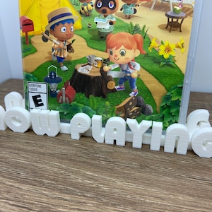May include: White plastic "now playing" sign with a video game case featuring a cartoon character on a green background. The game case has a "E for Everyone" rating.