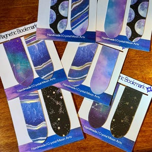 Op de afbeelding: Set van zes magnetische bladwijzers met een hemels thema. Elke bladwijzer heeft een ander ontwerp met een sterrenstelsel, sterren en manen in tinten paars, blauw en zwart.