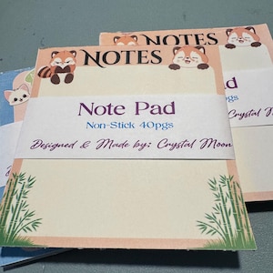 May include: Note pads with a light beige background and a pink border. The pads feature a red panda illustration at the top and bamboo illustrations at the bottom. The text "Note Pad" and "Non-Stick 40pgs" are visible.
