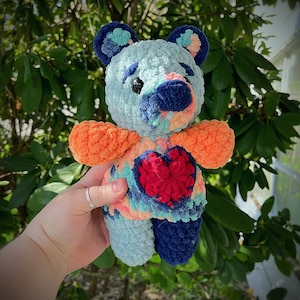 Puede incluir: Un oso de peluche tejido a crochet con un corazón en el pecho. El oso está hecho de hilos de diferentes colores, incluyendo azul, naranja y rojo. El corazón es rojo y tiene un contorno blanco.