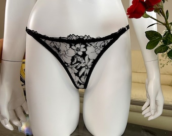 May include: Black lace thong underwear with a floral pattern displayed on a white mannequin. The underwear has a delicate, scalloped trim. A red rose is visible in the background. Sheet music is in the foreground.