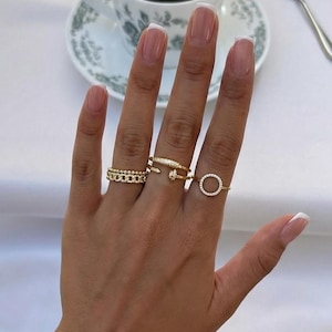 Peut inclure: Une main ornée de bagues en or, dont une de style chaîne, une bague avec un petit diamant et un cercle ouvert délicat. Une montre et des bracelets en argent sont également visibles. Une tasse de café est en arrière-plan.