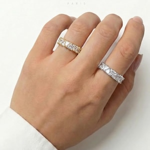 May include: Two eternity rings with square-cut gemstones are displayed on a hand. One ring is gold-toned, the other is silver-toned. The rings are set against a white background. The brand name "FRENCH COQUETTE PARIS" is visible.