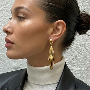 Può includere: Orecchini ovali allungati color oro con superficie strutturata e riflettente. Gli orecchini hanno un piccolo perno rotondo nella parte superiore. La modella indossa un dolcevita bianco e una giacca di pelle nera.