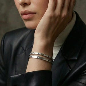 Può includere: Due braccialetti d'argento con decorazioni quadrate e a forma di stella sono indossati su un polso. I braccialetti sono indossati da una persona con una giacca di pelle nera e un dolcevita bianco. Lo sfondo è grigio neutro.
