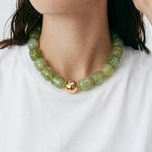 Peut inclure: Un collier de perles vertes avec un fermoir doré. Le collier est composé de grosses perles arrondies. Le modèle porte un t-shirt blanc et a deux petites boucles d'oreilles dorées.