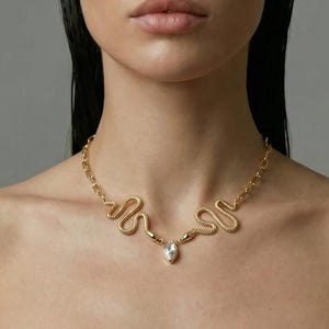 May include: A gold-toned necklace with a snake design, featuring a central pear-shaped gemstone. The necklace is worn against a white strapless top. The model has dark hair and is looking directly at the camera.