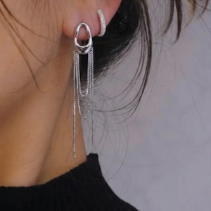 May include: Silver dangle earrings with an oval stud and multiple fine chain strands. A small, sparkly hoop earring is also visible. The earrings are shown against a blurred background.