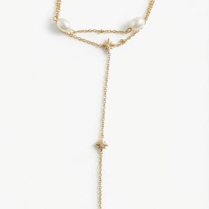 May include: A gold-toned lariat necklace featuring two white oval pearls and two star-shaped charms with small embedded stones. The delicate chain design includes a Y-shaped drop, with one star charm positioned at the junction and another further down.