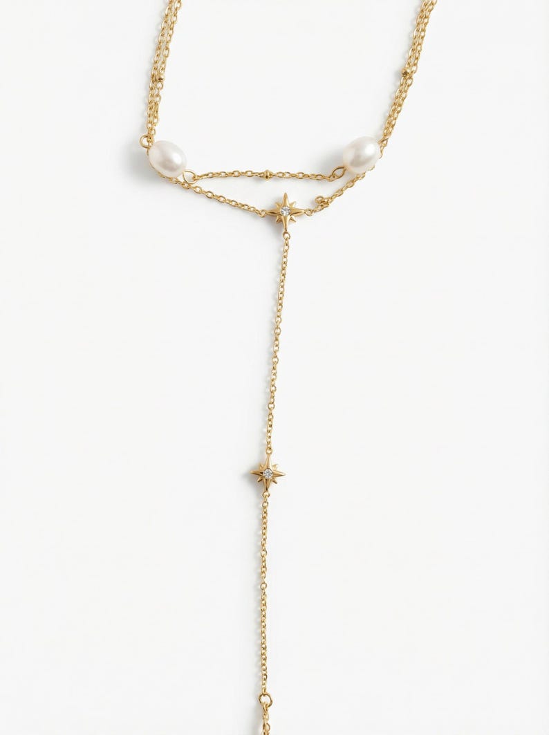 May include: A gold-toned lariat necklace featuring two white oval pearls and two star-shaped charms with small embedded stones. The delicate chain design includes a Y-shaped drop, with one star charm positioned at the junction and another further down.