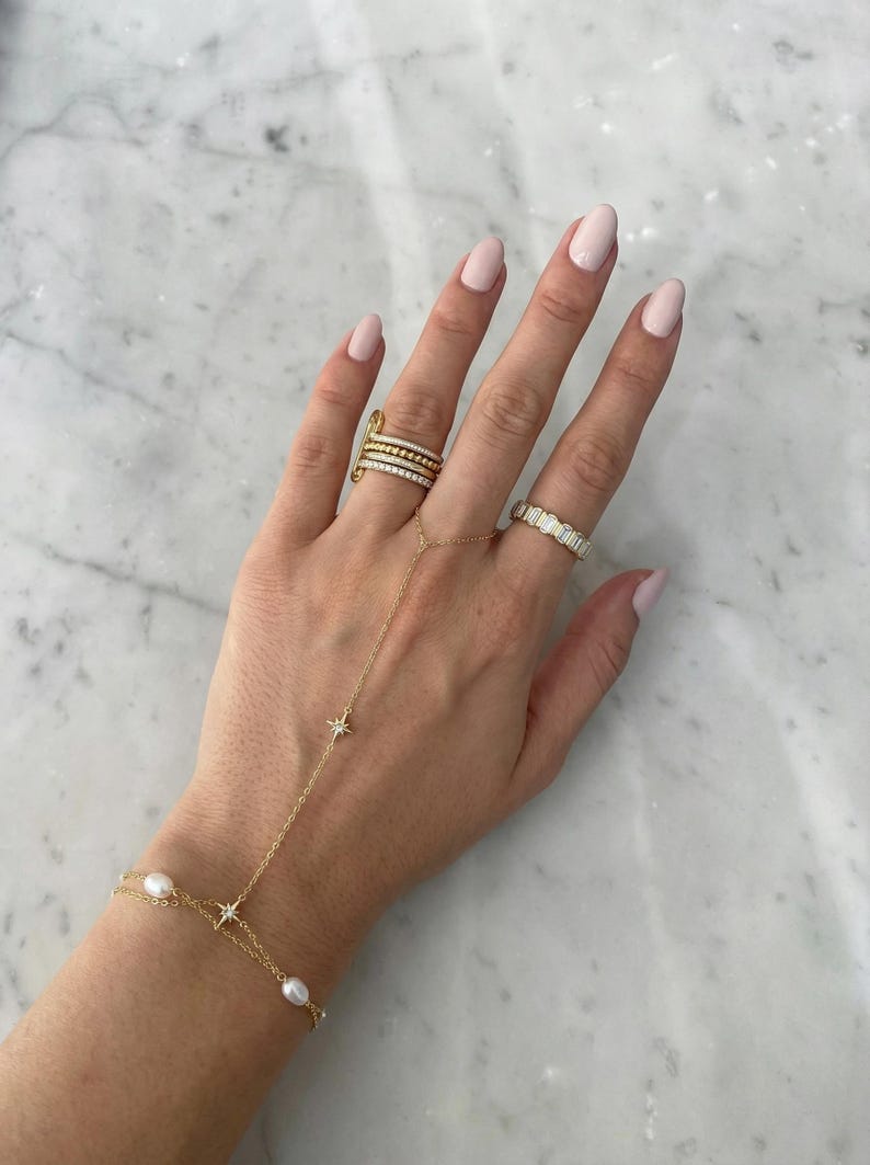 May include: A gold hand chain featuring small white pearls and star-shaped charms, connected to a ring. The hand also wears a stack of three thin gold rings on the middle finger and a gold band with rectangular stones on the ring finger.