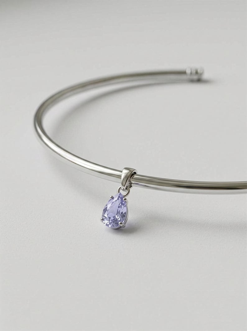 Puede incluir: Collar plateado con un colgante en forma de l&aacute;grima de color lila claro. El colgante cuelga de una barra curva de plata. La joya se muestra sobre un fondo blanco.