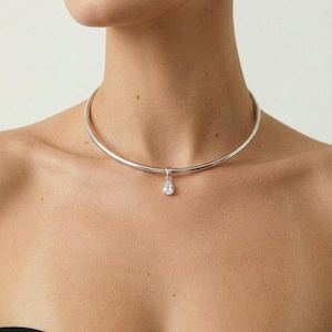 Puede incluir: Collar gargantilla plateado con un colgante en forma de l&aacute;grima. El collar es una banda de metal lisa y pulida. El colgante es una piedra preciosa transparente y brillante.