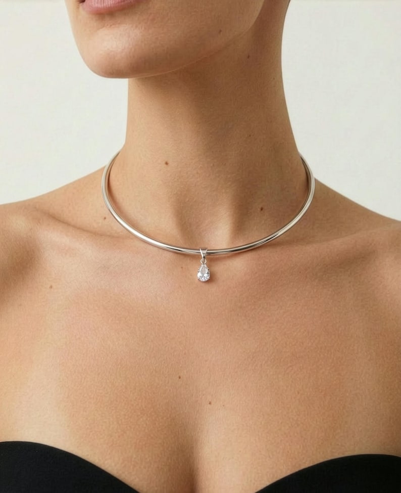 Puede incluir: Collar gargantilla plateado con un colgante en forma de l&aacute;grima. El collar es una banda de metal lisa y pulida. El colgante es una piedra preciosa transparente y brillante.