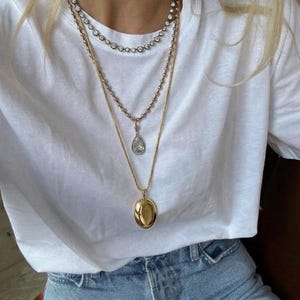 Op de afbeelding: Een gelaagde ketting set met een gouden medaillon, een druppelvormige hanger en een ketting met diamanten. De sieraden worden gedragen over een wit t-shirt en gecombineerd met een blauwe jeans.