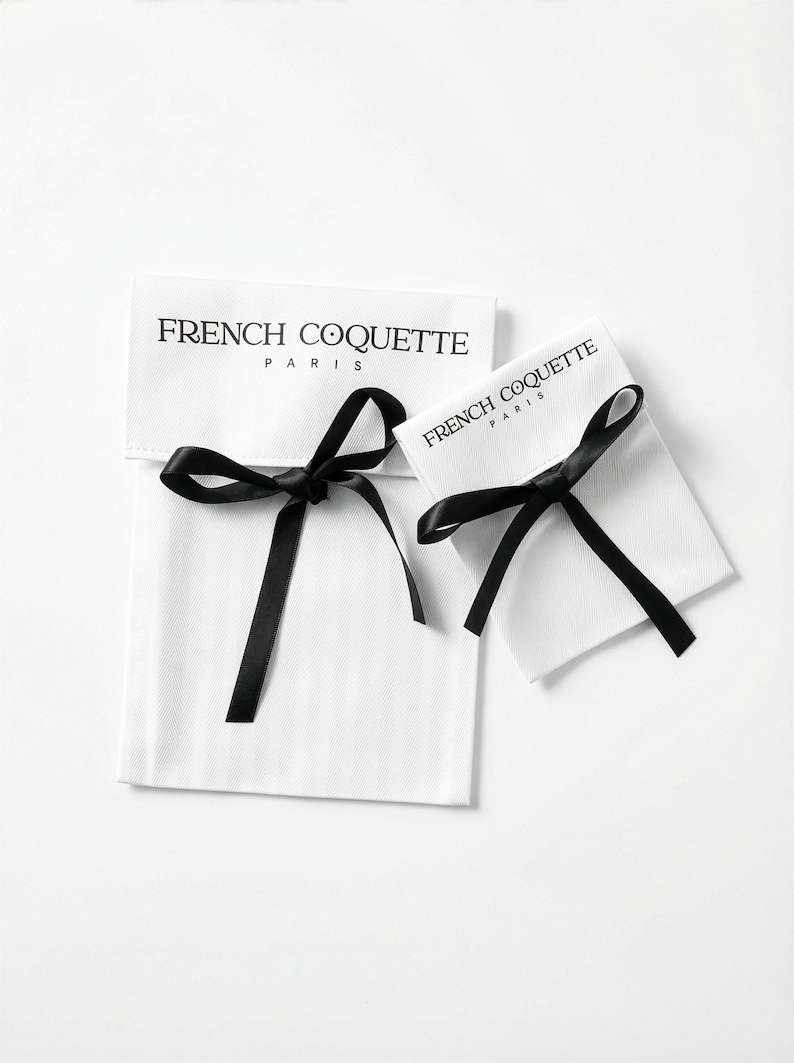 May include: Two white fabric pouches with black satin ribbon closures. The text "FRENCH COQUETTE PARIS" is printed in black on the front of each pouch. The pouches are likely for jewelry or small gifts.
