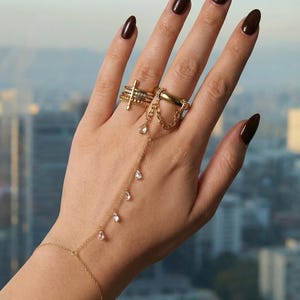 Puede incluir: Una mano adornada con anillos de oro y una cadena de mano. Los anillos incluyen una pila de bandas, un anillo con un diseño de cruz y una banda gruesa. La cadena de mano presenta gemas colgantes en forma de lágrima.
