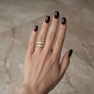 Peut inclure: Une main avec du vernis à ongles bordeaux foncé, présentant deux bagues en or. L'une des bagues comporte une rangée de pierres rectangulaires, l'autre des pierres ovales. Les bagues sont posées sur un fond neutre.